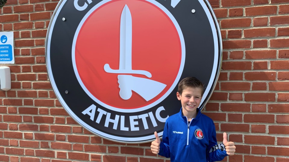 Harrison Wickers with his thumbs up standing next to Charlton Athletic logo